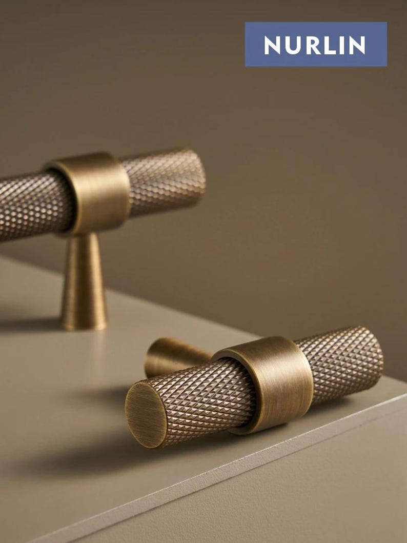 May include: Close-up of two antique brass cabinet pulls. The pulls have a cylindrical, textured handle with a diamond pattern and are attached to a round base. The word "NURLIN" is printed on a blue rectangle in the background.