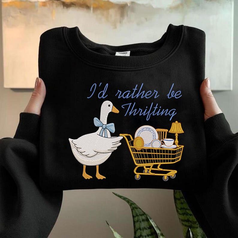 May include: Black sweatshirt with the phrase "I'd rather be Thrifting" in blue script. A white goose with a blue bow tie is next to a shopping cart filled with dishes and a lamp.
