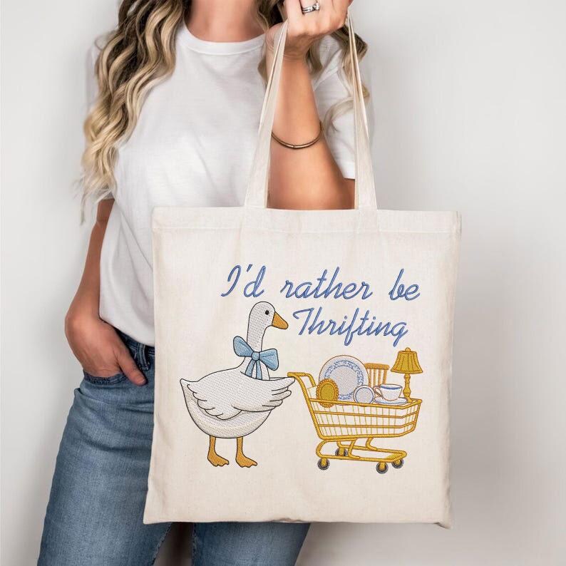 May include: Cream-colored tote bag with the phrase "I'd rather be thrifting" in blue script. The design features a goose wearing a blue bow and pushing a shopping cart filled with dishes and a lamp.