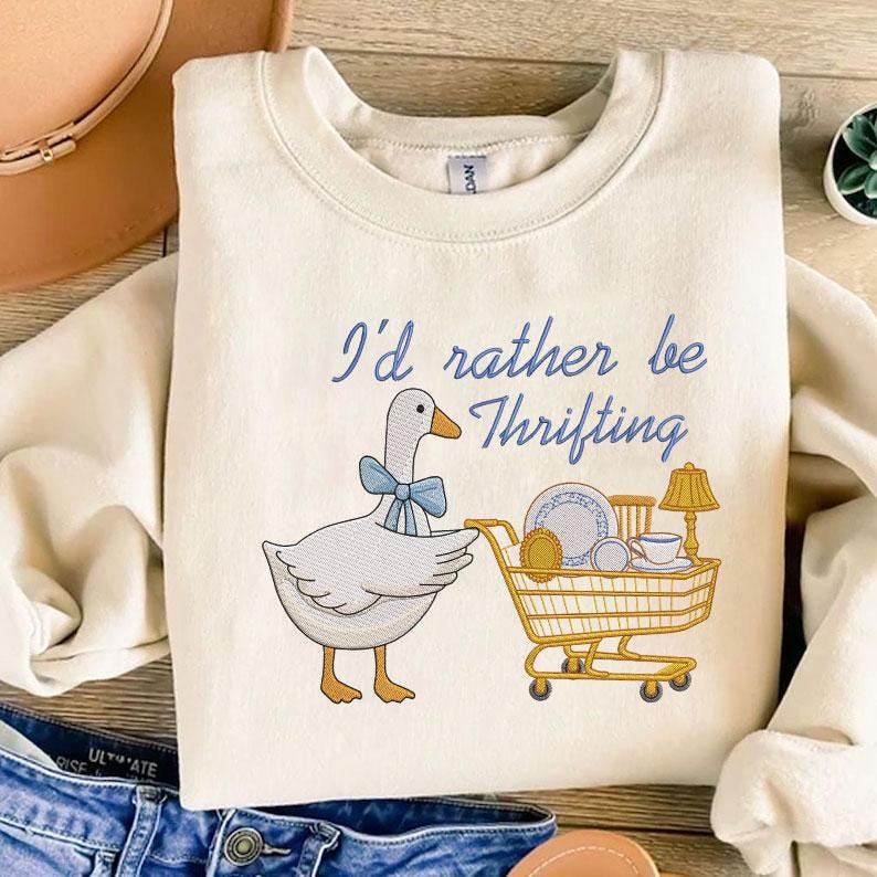 May include: Cream-colored sweatshirt with the embroidered phrase "I'd rather be Thrifting." A cartoon goose with a blue bow tie pushes a shopping cart filled with vintage items. The design includes a lamp, teacup, and plates.
