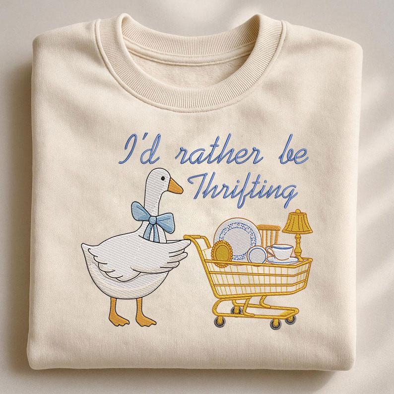 May include: Cream-colored sweatshirt with the embroidered phrase "I'd rather be Thrifting." A cartoon goose with a blue bow tie pulls a shopping cart filled with vintage items. The design features a lamp, teacup, and plates.
