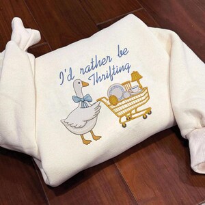 May include: Cream-colored sweatshirt with the embroidered phrase "I'd rather be thrifting." A cartoon goose with a blue bow tie pulls a yellow shopping cart filled with dishes and a lamp. The sweatshirt has ribbed cuffs and a folded neckline.