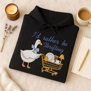 May include: Black hooded sweatshirt with the embroidered phrase "I'd rather be Thrifting." A white goose with a blue bow tie pulls a shopping cart filled with thrifted items. A lit candle, coffee cup, and notebook are in the background.