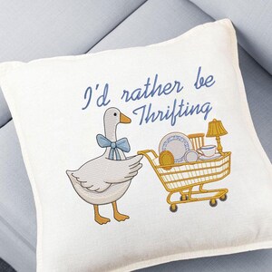 May include: Cream-colored decorative pillow with an embroidered design. A cartoon goose with a blue bow tie is pushing a yellow shopping cart filled with vintage items. The text "I'd rather be Thrifting" is embroidered in blue.