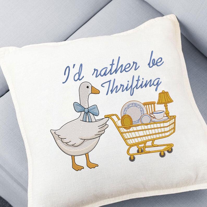May include: Cream-colored decorative pillow with an embroidered design. A cartoon goose with a blue bow tie is pushing a yellow shopping cart filled with vintage items. The text "I'd rather be Thrifting" is embroidered in blue.
