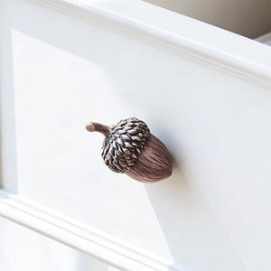 Puede incluir: Primer plano de un cajón blanco con otro cajón abierto encima. El cajón presenta un tirador en forma de bellota de color bronce. El tirador de bellota tiene una tapa texturizada y un cuerpo liso y redondeado.