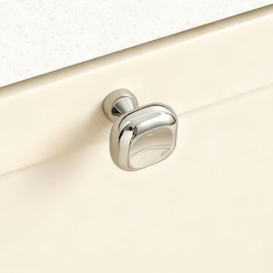 May include: A polished chrome cabinet knob with a square face and rounded edges. The knob is mounted on a cream-colored cabinet door. The background features a white countertop.