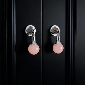 以下が含まれることがあります： 光沢のあるクロム仕上げと、淡いピンク色の球形宝石が付いた2つのキャビネットプル。プルは黒いキャビネットドアに取り付けられています。プルはモダンでエレガントなデザインです。