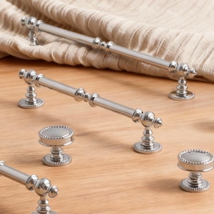 Könnte beinhalten: Eine Sammlung von silberfarbenen Schrankbeschlägen, darunter lange Stangengriffe und runde Knöpfe. Die Beschläge weisen ein detailliertes, perlenbesetztes Design und eine klassische, elegante Ästhetik auf. Die Teile sind auf einer hellen Holzoberfläche angeordnet.