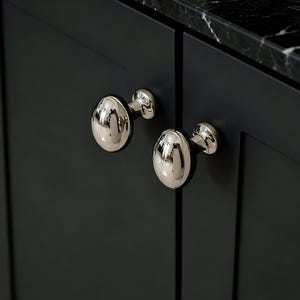 Könnte beinhalten: Nahaufnahme von zwei polierten Nickel-Schrankknöpfen mit ovaler Form. Die Knöpfe sind an einer dunkelgrauen Schranktür angebracht. Die Arbeitsplatte ist schwarz mit weißen Adern.