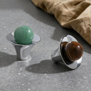 May include: Two decorative bottle stoppers. One features a polished green stone sphere, the other a brown stone sphere. Both are set in a shiny, silver-colored metal base with a flared design. The background includes a tan fabric.