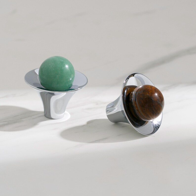 K&ouml;nnte beinhalten: Zwei dekorative Objekte mit polierter Chromoberfl&auml;che. Eines h&auml;lt eine gr&uuml;ne Kugel, das andere eine braune Kugel. Die Objekte haben einen ausgestellten Sockel und eine kreisf&ouml;rmige Oberseite, was eine moderne, minimalistische &Auml;sthetik schafft.