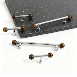 Könnte beinhalten: Eine Sammlung von Schrankgriffen mit modernem Design. Jeder Griff hat eine polierte Chromstange mit einem dekorativen, runden Tigeraugenstein an jedem Ende. Die Griffe sind in verschiedenen Größen erhältlich, der längste misst etwa 30 cm.