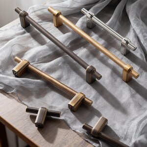 May include: A collection of cabinet pulls in various metallic finishes, including bronze, gold, and silver. The pulls have a rectangular base with a cylindrical bar. The hardware is displayed on a gray fabric surface.