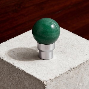 Op de afbeelding: Een gepolijste, groene edelsteenbol gemonteerd op een zilverkleurige metalen basis. De bol heeft een levendige groene tint en staat op een cilindrische basis. Het object wordt getoond op een betonnen blok.