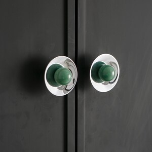 K&ouml;nnte beinhalten: Nahaufnahme von zwei Schrankt&uuml;rkn&ouml;pfen. Jeder Knopf hat eine polierte Chrombasis mit kreisf&ouml;rmigem Design und einen abgerundeten, gr&uuml;nen Mittelteil. Die Kn&ouml;pfe sind an einer dunkelgrauen Schrankt&uuml;r befestigt.