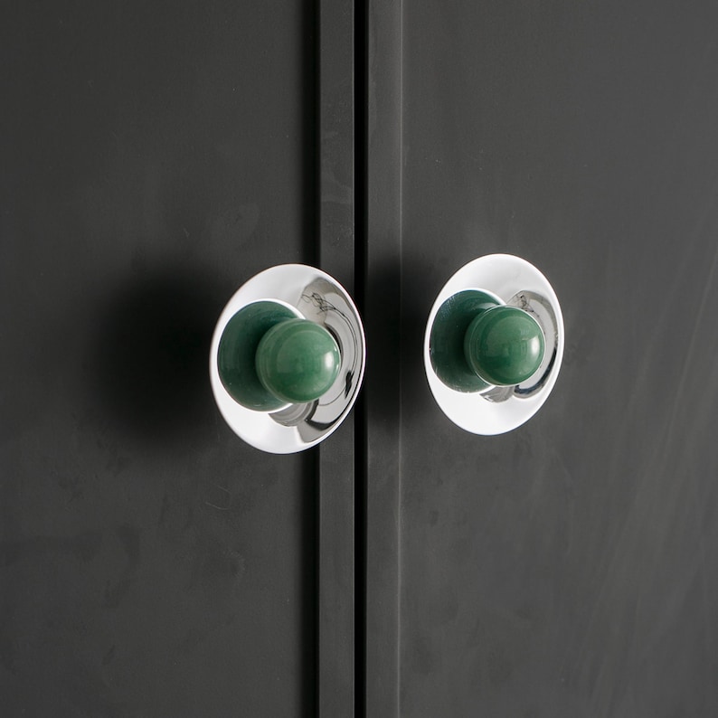 K&ouml;nnte beinhalten: Nahaufnahme von zwei Schrankt&uuml;rkn&ouml;pfen. Jeder Knopf hat eine polierte Chrombasis mit kreisf&ouml;rmigem Design und einen abgerundeten, gr&uuml;nen Mittelteil. Die Kn&ouml;pfe sind an einer dunkelgrauen Schrankt&uuml;r befestigt.