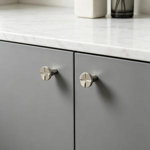 May include: Two circular cabinet knobs with a polished silver finish and a cross-shaped indentation on the face. The knobs are mounted on two adjacent matte gray cabinet doors, each featuring a central stem connecting to the circular head.