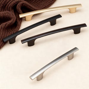 May include: A collection of cabinet pulls in various metallic finishes. The pulls are in gold, black, dark brown, and silver. Each pull has a curved bar design with two rounded feet.