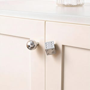 Puede incluir: Dos tiradores de armario. Uno es un tirador redondo con acabado cromado. El otro es un tirador con acabado cromado en forma de cubo diseñado para parecer un dado, con puntos visibles. Ambos tiradores están montados en una puerta de armario color crema.