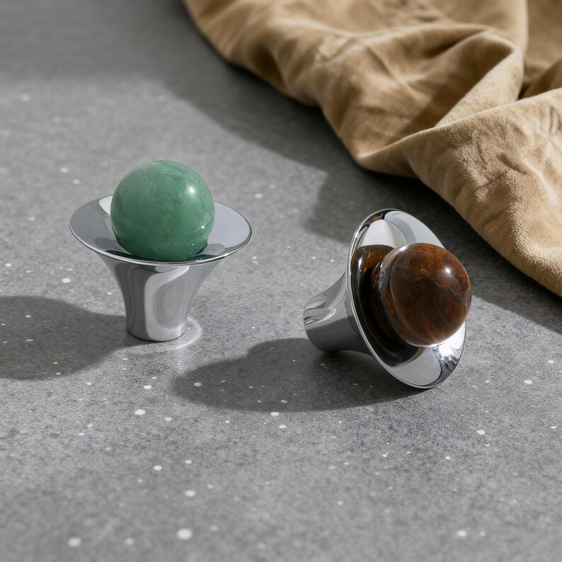 May include: Two decorative bottle stoppers. One features a polished green stone sphere, the other a brown stone sphere. Both are set in a shiny, silver-colored metal base with a flared design. The background includes a tan fabric.