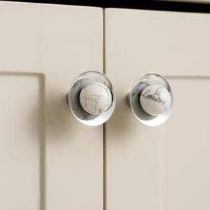 K&ouml;nnte beinhalten: Zwei Schrankkn&ouml;pfe mit klarer, kreisf&ouml;rmiger Basis und einer wei&szlig;en, marmor&auml;hnlichen Kugel in der Mitte. Sie sind an einer hellbeigen Schrankt&uuml;r montiert und bieten eine moderne und elegante &Auml;sthetik.