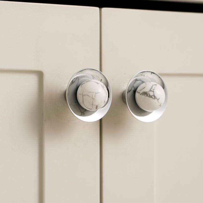 K&ouml;nnte beinhalten: Zwei Schrankkn&ouml;pfe mit klarer, kreisf&ouml;rmiger Basis und einer wei&szlig;en, marmor&auml;hnlichen Kugel in der Mitte. Sie sind an einer hellbeigen Schrankt&uuml;r montiert und bieten eine moderne und elegante &Auml;sthetik.