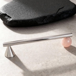 Könnte beinhalten: Ein moderner, minimalistischer Schrankgriff. Der Griff besteht aus poliertem Metall mit einer zylindrischen Stange und einem konischen Sockel. Am gegenüberliegenden Ende der Stange befindet sich eine rosa Kugel. Der Griff ist vor neutralem Hintergrund.