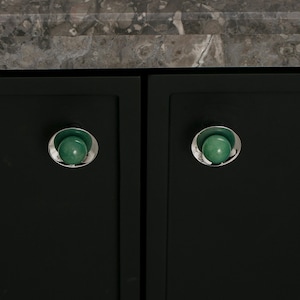 K&ouml;nnte beinhalten: Nahaufnahme von zwei dunkelgr&uuml;nen Schrankt&uuml;ren mit passender Hardware. Jede T&uuml;r hat einen silberfarbenen Knopf mit einem gr&uuml;nen, kugelf&ouml;rmigen Zentrum. Die Kn&ouml;pfe sind vor einem dunklen Hintergrund platziert, mit einer Marmorarbeitsplatte dar&uuml;ber.