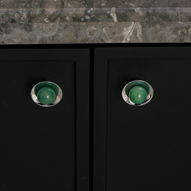 K&ouml;nnte beinhalten: Nahaufnahme von zwei dunkelgr&uuml;nen Schrankt&uuml;ren mit passender Hardware. Jede T&uuml;r hat einen silberfarbenen Knopf mit einem gr&uuml;nen, kugelf&ouml;rmigen Zentrum. Die Kn&ouml;pfe sind vor einem dunklen Hintergrund platziert, mit einer Marmorarbeitsplatte dar&uuml;ber.