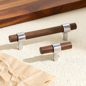 Könnte beinhalten: Zwei Schrankgriffe mit einem dunkelbraunen Holzfass und einem silbernen Metallring und Sockel. Ein Griff ist länger als der andere. Im Hintergrund eine dunkelbraune Holzoberfläche.