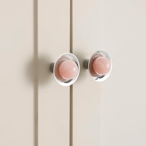 K&ouml;nnte beinhalten: Zwei Schrankkn&ouml;pfe mit einem hellrosa, kugelf&ouml;rmigen Mittelteil und einem silbernen, kreisf&ouml;rmigen Au&szlig;enring. Die Kn&ouml;pfe sind an einer hellbeige Schrankt&uuml;r befestigt. Die Kn&ouml;pfe haben ein modernes, minimalistisches Design.