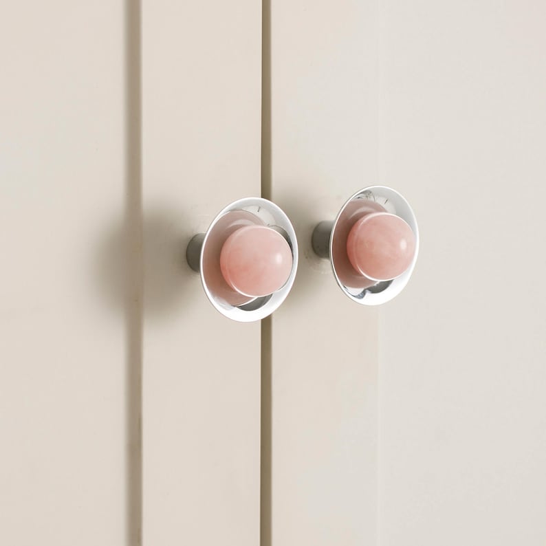 K&ouml;nnte beinhalten: Zwei Schrankkn&ouml;pfe mit einem hellrosa, kugelf&ouml;rmigen Mittelteil und einem silbernen, kreisf&ouml;rmigen Au&szlig;enring. Die Kn&ouml;pfe sind an einer hellbeige Schrankt&uuml;r befestigt. Die Kn&ouml;pfe haben ein modernes, minimalistisches Design.