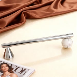 Może przedstawiać: Polerowany chromowany drążek na ręczniki z białą kulą z marmuru na jednym końcu i chromowanym stożkiem na drugim. Drążek jest ustawiony na tle brązowej satynowej tkaniny i magazynu.