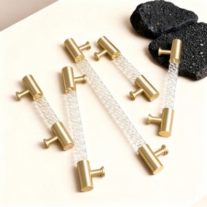 Peut inclure: Six poignées de meubles avec un centre en verre torsadé transparent et des extrémités en métal doré. Les poignées sont disposées sur un fond clair, mettant en valeur leur design et leurs détails. Quincaillerie décorative.