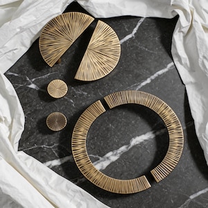 Op de afbeelding: Een verzameling messingkleurige hardware, waaronder twee halve cirkelvormige handgrepen, twee kleine ronde knoppen en een gesegmenteerde ring. De hardware heeft een gestructureerd, radiaal ontwerp. De items zijn gerangschikt op een donker, gemarmerd oppervlak met witte stof op de achtergrond.