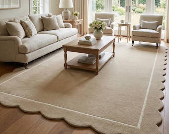 May include: A beige rectangular area rug with a scalloped edge and a cream-colored border. The rug is placed on a hardwood floor in a living room setting, with a sofa, chairs, and a coffee table.