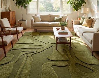 May include: A living room featuring a large, olive green area rug with an abstract, raised design. The rug is the focal point, complemented by neutral-toned sofas, wooden chairs, and a coffee table. Large potted plants and natural light enhance the room's aesthetic.