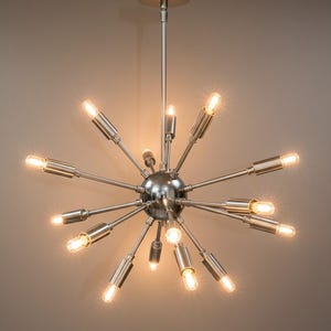 Könnte beinhalten: Eine moderne Sputnik-Chandelier in Silberoptik mit mehreren Armen, die von einer zentralen Kugel ausgehen. Jeder Arm trägt eine kleine, leuchtende Glühbirne, die einen Sternenstrahleffekt erzeugt. Die Leuchte wird an einer Deckenhalterung aufgehängt.