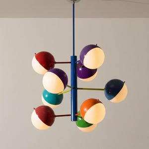 Può includere: Un lampadario moderno con una colonna centrale e bracci blu, che supporta più luci sferiche. Le sfere sono divise in sezioni colorate di rosso, viola, turchese, arancione, verde e blu navy, con una metà bianca illuminata. Fissaggio a soffitto cromato.