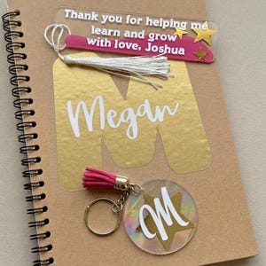 May include: A brown spiral-bound notebook with a gold letter 'M' and a clear bookmark with the text 'Thank you for helping me learn and grow with love, Joshua'. A keychain with a pink tassel and the letter 'M' is also visible.