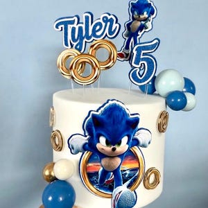 Könnte beinhalten: Eine weiße zylindrische Torte mit Sonic-the-Hedgehog-Motiven, verziert mit einem blauen Glitzer-Namensaufleger Tyler, einer blauen Glitzer-5, goldenen Ringakzenten sowie kleinen blauen und weißen kugelförmigen Ballondekorationen.