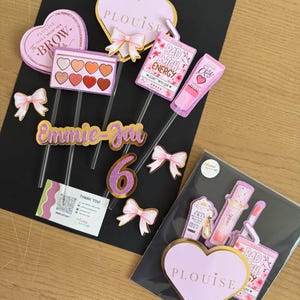 Op de afbeelding: Een verzameling roze en gouden taarttoppers en decoraties. Inclusief hartvormige toppers met de tekst "PLOUISE", strikken en make-up-thema ontwerpen. Bevat ook de naam "Emmie-Jai" en het nummer "6".