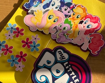 May include: A collection of My Little Pony stickers and cutouts on a yellow background. The stickers feature various colorful ponies and a purple dragon. Additional flower stickers are also present, along with a My Little Pony logo sticker.