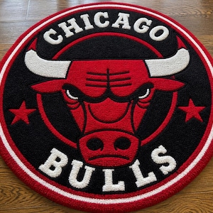 May include: A round rug featuring the Chicago Bulls logo. The rug has a red bull head in the center, surrounded by a black background. The words "CHICAGO" and "BULLS" are in white, with red stars on either side of the bull's head. The rug is trimmed with a white and red border.