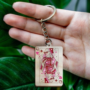 May include: Wooden keychain featuring a playing card design of the Queen of Hearts. The card has a pink and red color scheme with a stylized illustration of a woman wearing sunglasses. The keychain includes a metal ring and chain.