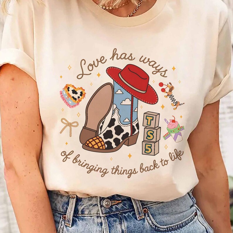 May include: A cream-colored t-shirt featuring a graphic print with a red cowboy hat, a cow-print boot, a stack of blocks labeled T S 5, a heart, a bow, and a toy dog. The text reads Love has ways of bringing things back to life.