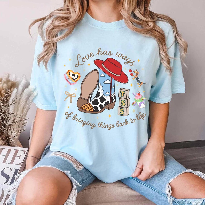 May include: A light blue t-shirt featuring a graphic print of a cowboy boot with a cow print pattern, a red hat, three stacked alphabet blocks with the letters T, S, and 5, and the text Love has ways of bringing things back to life.