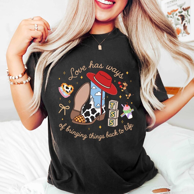 May include: A black t-shirt featuring a graphic print with the text "Love has ways of bringing things back to life." The design includes a cowboy boot, a red hat, a heart-shaped object, a toy dog, alphabet blocks with "T S 5," and a drink cup.