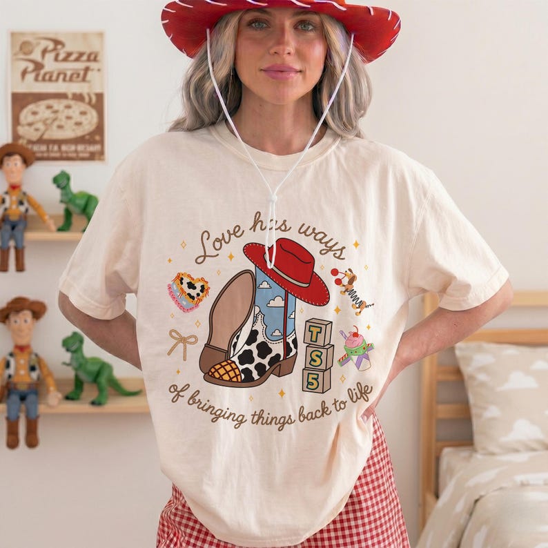 May include: A cream-colored t-shirt featuring a graphic print of a red cowboy hat, a cow-print boot, alphabet blocks, and the text 'Love has ways of bringing things back to life'. The shirt is worn with a red cowboy hat with a white chin strap.